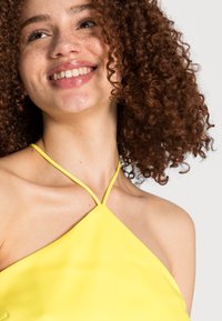Top amarelo de gola halter com um design triangular e alças finas, apresentando uma textura suave e um estilo minimalista, destacando a linha do pescoço.