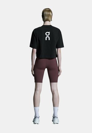 Personne debout de dos portant un t-shirt court noir avec un logo blanc, un short de cyclisme bordeaux, des chaussettes blanches et des baskets blanches.