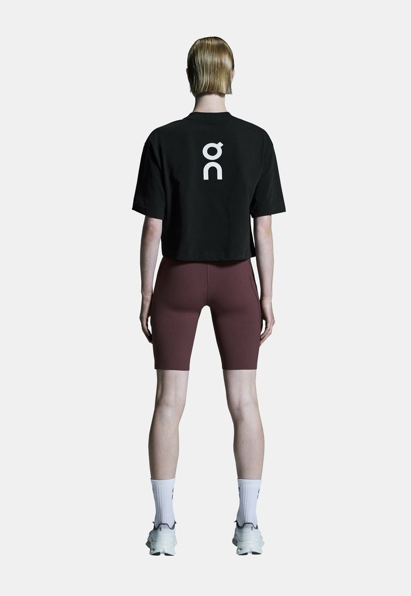 Personne debout de dos portant un t-shirt court noir avec un logo blanc, un short de cyclisme bordeaux, des chaussettes blanches et des baskets blanches.
