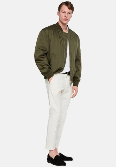 Giacca bomber verde oliva con zip frontale, abbinata a pantaloni bianchi a sigaretta e mocassini neri. Presenta tasche laterali e una texture liscia.