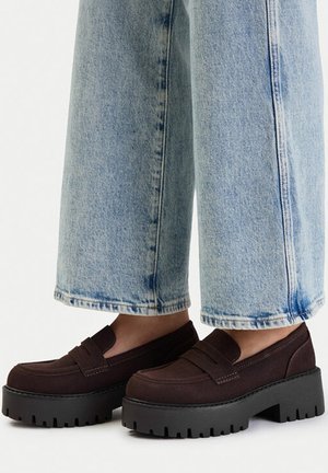 Persona con vaqueros de pierna ancha azul claro y mocasines plataforma gruesos de color marrón oscuro con suelas gruesas estriadas.