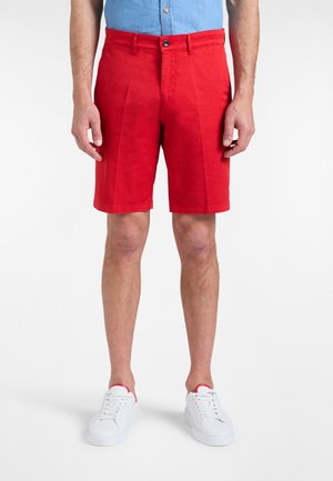 Muškarac u svijetlo crvenim kratkim hlačama do koljena, bijelim tenisicama i svijetloplavoj košulji uvučenoj u hlače, stoji na bijeloj pozadini.