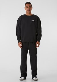 Hanorac negru cu mâneci lungi, manșete rib, și guler rotund. Asortat cu pantaloni de trening negri și adidași gri.