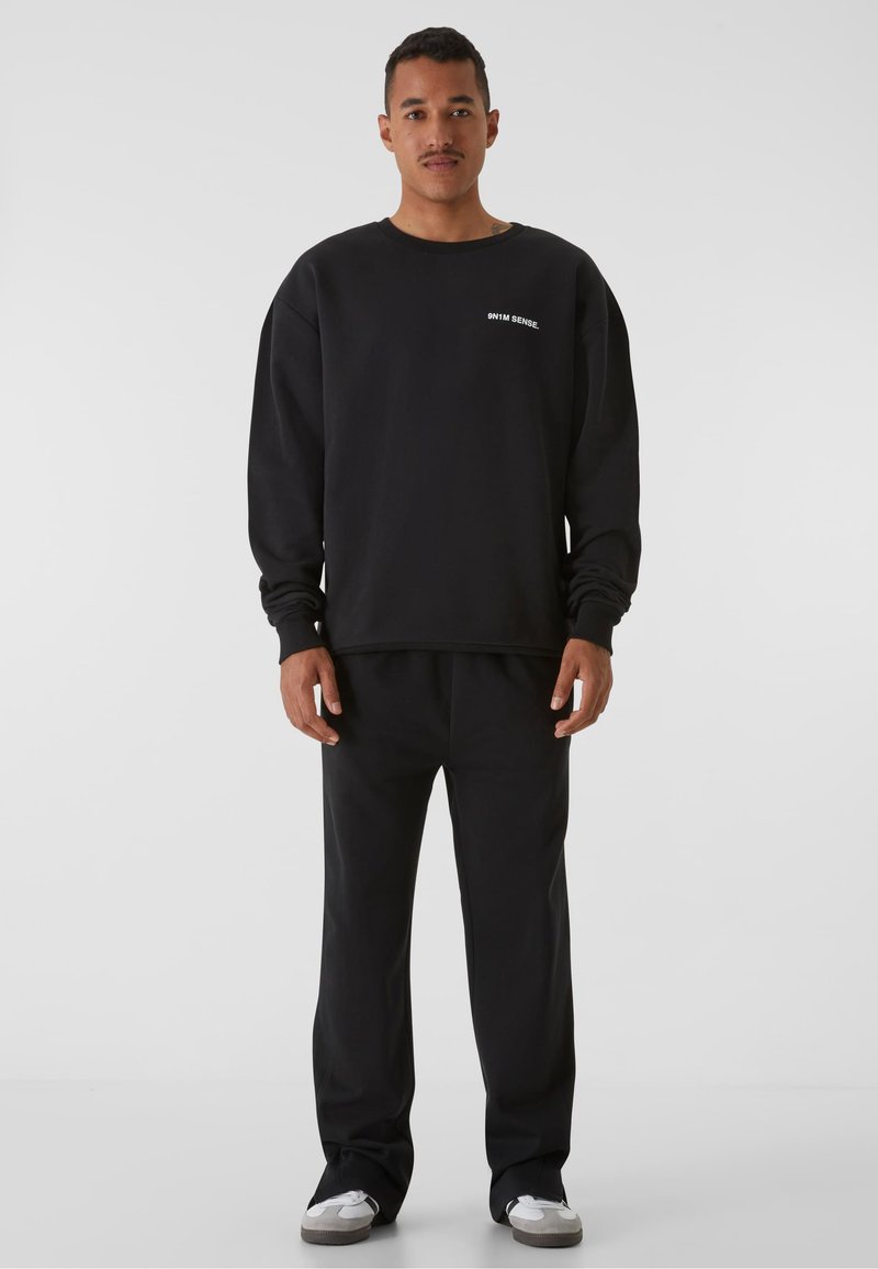 Hanorac negru cu mâneci lungi, manșete rib, și guler rotund. Asortat cu pantaloni de trening negri și adidași gri.