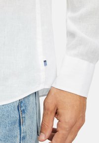 Weißes Leinenhemd mit einem Manschette und Seitennähten. Besitzt einen blauen gestreiften Akzent am unteren Saum. Getragen mit hellblauen Jeans.