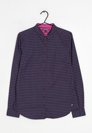 Chemise bleu marine à manches longues avec boutons, motif géométrique à pois roses, col intérieur rose, sur un cintre noir contre un fond blanc.