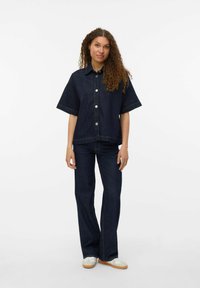 Mörk denim oversized skjorta med korta ärmar, framknäppning och matchande mörka denimflared byxor. Snygga vita sneakers fulländar looken.