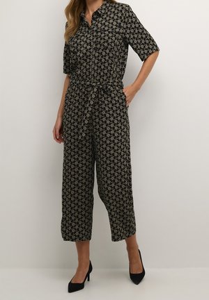 Mono negro con estampado floral dorado, mangas cortas, escote con cuello, cintura con cinturón y piernas anchas recortadas. Tacones altos negros complementan el conjunto.