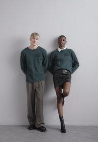 Two models stand against a plain background. The male wears a green ribbed sweater and gray trousers. The female wears a green sweater over a white collared shirt and a black leather skirt.