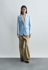 Γυναίκα που στέκεται μπροστά σε γκρι τοίχο, φοράει ανοιχτό μπλε σακάκι, μπεζ παντελόνια με πλατιά πόδια και διάτρητα μοτίβα, και μαύρα παπούτσια.