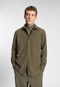 Olijfgroene langemouwen blouse met kraag, knoopsluiting aan de voorkant en subtiele textuur. Draagt over een bijpassende coltrui, met recht model.