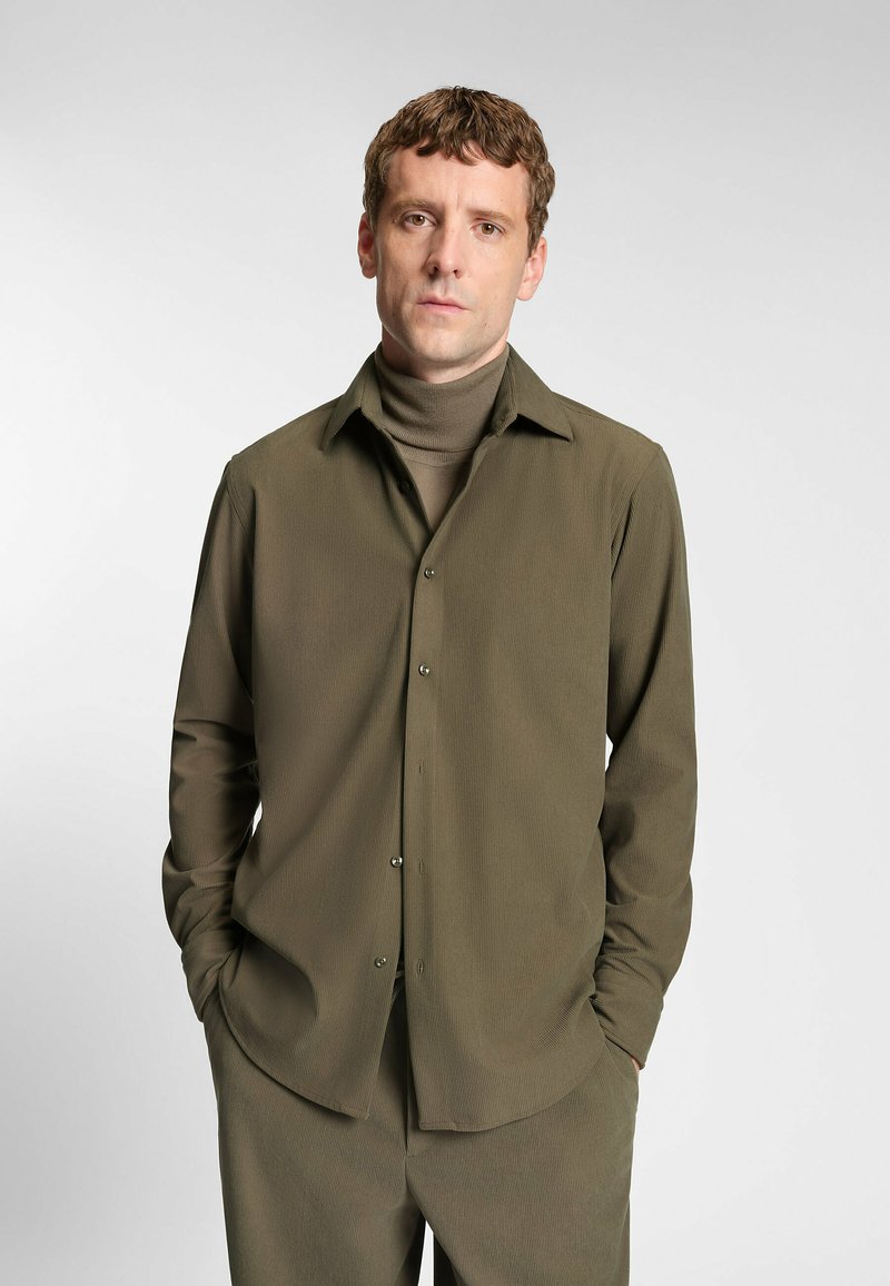 Olijfgroene langemouwen blouse met kraag, knoopsluiting aan de voorkant en subtiele textuur. Draagt over een bijpassende coltrui, met recht model.