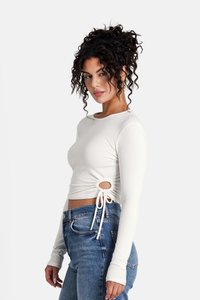 Maglietta grigia chiara a maniche lunghe con una texture a coste, presenta un'apertura laterale e una cintura regolabile. Abbinata a jeans blue a vita alta.