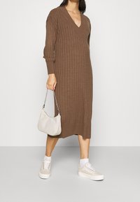Donna che indossa un vestito midi a coste marrone, calze bianche, sneakers beige e tiene in mano una piccola borsa bianca con una tracolla in argento.