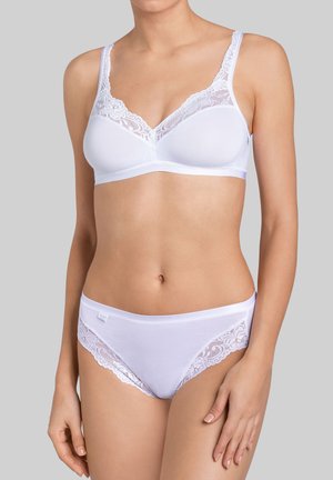 Nainen, jolla on valkoinen pitsireunainen rintaliivit ja yhteen sopivat alushousut, seisoo yksinkertaisen harmaan taustan edessä.