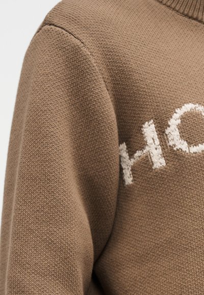 Κοντινό πλάνο ενός καφέ πλεκτού πουλόβερ με μερικό λευκό υφασμάτινο λογότυπο στο στήθος και ριμπ γύρο λαιμόκοψη.