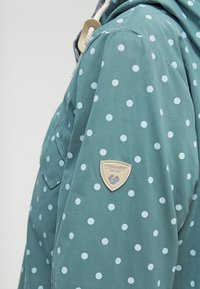 Niebieskozielona kurtka przeciwdeszczowa w białe grochy, z kapturem i naszywką na rękawie identyfikującą markę. Gładka faktura tkaniny.