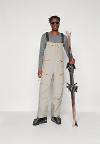 Béžové snehové overalové nohavice s oranžovými zipsami a viacerými vreckami, nosené cez sivé tričko s dlhým rukávom. Osoba drž�í pár lyží.