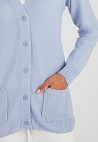 Cardigan lavorato a maglia azzurro chiaro con scollatura a V, bottoni frontali, polsini a coste e due tasche frontali. Il tessuto è morbido e testurizzato.