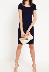 Tmavě modré přiléhavé šaty s krátkými rukávy, hladká textura. Modelka drží béžovou obálkovou kabelku s klopou, v kombinaci s béžovými sandály na podpatku.