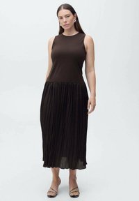 Rochie maro fără mâneci, cu un top aderent și fustă pliată, având o textură fină și un design până la glezne. Sandale negre simple completează ținuta.