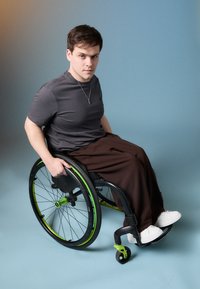Fauteuil roulant avec cadre noir, accents verts et grandes roues ; personne en chemise grise et pantalon marron, chaussures blanches, assise avec une expression neutre.