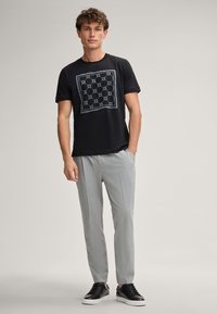 Zwarte T-shirt met een vierkant grafisch ontwerp in grijs, gecombineerd met lichtgrijze geblokte broek en zwarte sneakers met witte zolen.