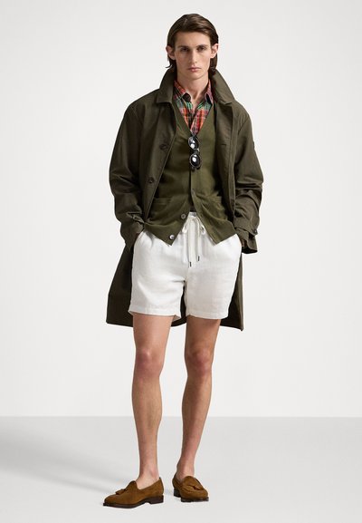 Un pardesiu verde peste o vestă verde închis, cămașă cu carouri, pantaloni scurți albi și mocasini maro. Accesoriile includ ochelari de soare negri.