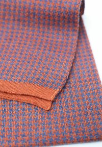 Knitted fabric in orange and blue houndstooth pattern, featuring a smooth texture with a folded edge and a plain hem.