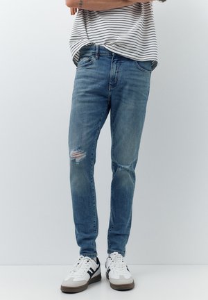 Persoană care poartă blugi skinny albaștri cu rupturi, adidași albi cu dungi negre și o cămașă albă cu dungi orizontale negre.