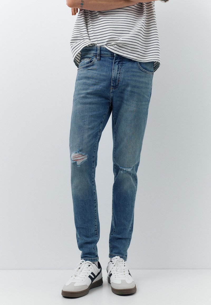 Personne portant un jean skinny bleu déchiré, des baskets blanches avec des rayures noires, et une chemise blanche à rayures horizontales noires.