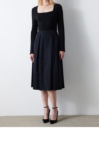 Top nero a maniche lunghe con scollo quadrato abbinato a una gonna midi plissettata scura. Il modello indossa tacchi con cinturino alla caviglia neri. Texture complessiva liscia.