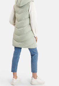 Jasnozielona pikowana kamizelka z kapturem, ozdobiona wzorem w jodełkę i pikowanym designem. Noszona na białym swetrze i niebieskich dżinsach.