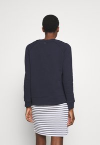 Námořnická modrá mikina s žebrovanými manžetami a spodním lemem, má raglánové rukávy a nenápadnou značku na zadní straně. Spárována s pruhovanými šaty.