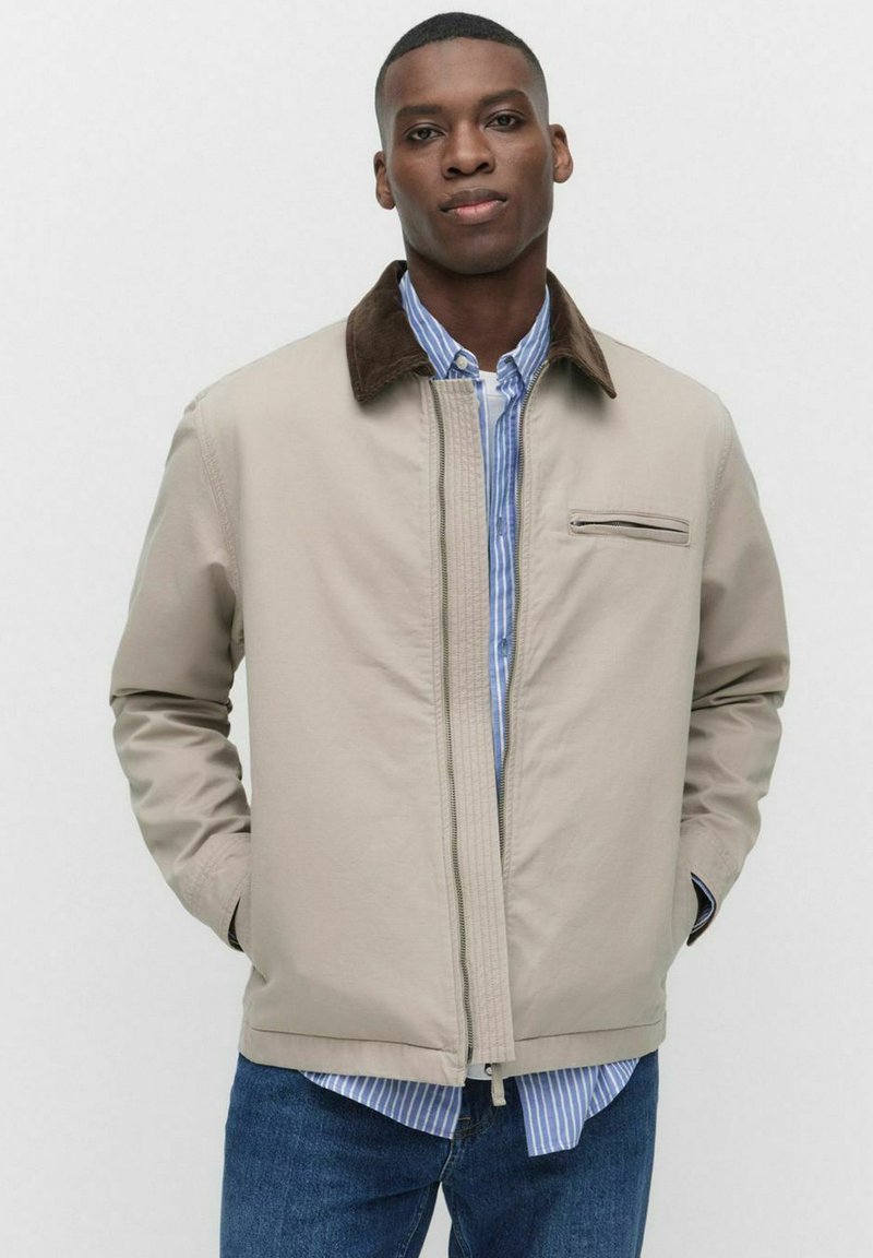 Veste beige clair avec un col marron, fermeture éclair à l'avant et une poche poitrine. Portée par-dessus une chemise rayée bleu et blanc, associée à un jean bleu.