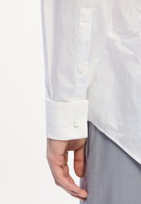 Poignet de chemise blanche avec une texture lisse, mettant en avant un bouton unique et un éclat subtil. Tissu gris visible en dessous du poignet.