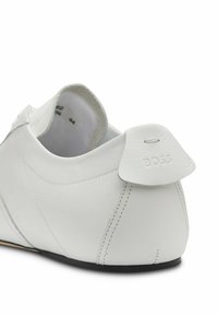 Chaussure basse en cuir blanc avec détails cousus, bord de la semelle noir et "BOSS" embossé sur une languette arrière.