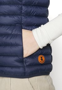 Piumino smanicato blu navy con trapuntatura orizzontale, caratterizzato da una toppa arancione. Lo smanicato ha una tasca laterale e una texture liscia e lucida.