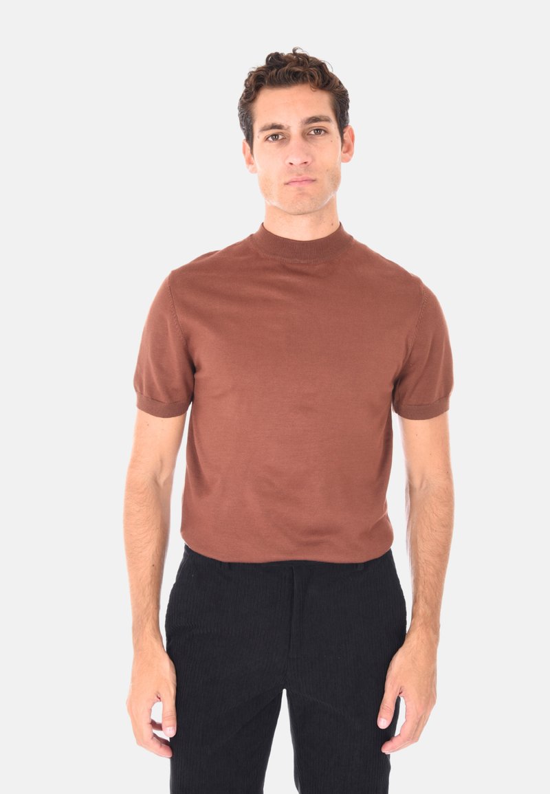 Uomo con capelli corti e ricci, che indossa una maglietta a maniche corte marrone e pantaloni neri, in piedi contro uno sfondo chiaro e semplice.
