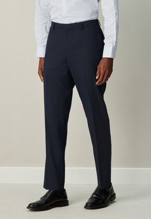 Mand iført skræddersyede mørkeblå bukser, sorte læder monk strap sko og en lyseblå stribet skjorte, der står op ad en enkel væg.