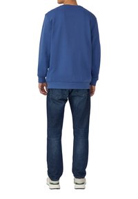 Una persona che indossa una felpa a maniche lunghe blu, jeans blu scuro e sneakers bianche, in piedi e con la schiena rivolta verso una superficie bianca.