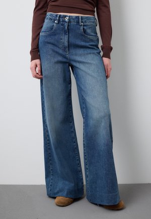 Weit geschnittene blaue Jeans aus Denim, getragen mit braunem Langarmshirt und hellbraunen Schuhen, stehend auf grauem Boden vor weißer Wand.