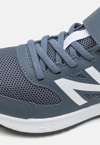 Šedá sportovní bota s síťovaným svrškem, polstrovaným jazykem a bílým logem. Obsahuje texturované akcenty a gumovou podrážku pro trakci.