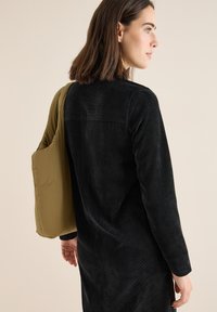 Abito in velluto a coste nero con maniche lunghe, caratterizzato da uno yoke posteriore e una borsa a tracolla verde oliva imbottita appesa sulla spalla.