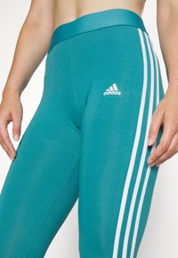 Teal leggings med bred midja, med tre vita vertikala ränder på sidan och ett Adidas-logotyp framtill. Slät textur.