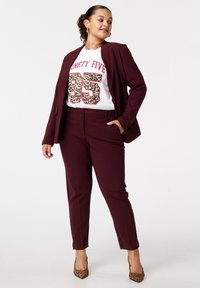Bordeaux op maat gemaakte kostuum met slim-fit broek en blazer, gedragen over een witte graphic tee met een luipaardprint nummer "95," gecombineerd met dierenprint hakken.