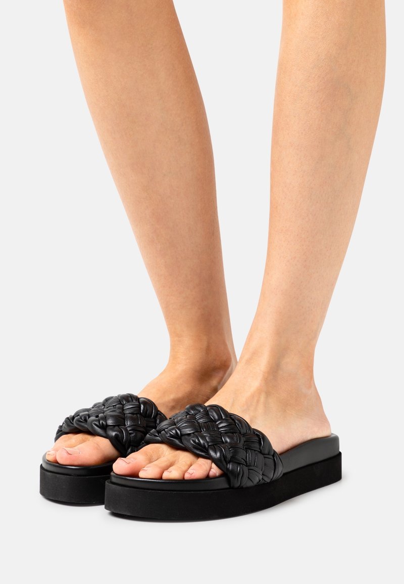 Sandales plates noires tissées avec une semelle épaisse et une finition texturée. La tige présente un design tressé, fabriqué en matériau doux semblable au cuir.