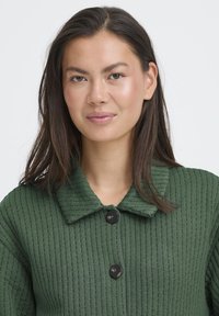 Grøn struktureret jakke med et ribbet mønster, der har en krave og tre sorte knapper. Stoffet ser blødt og letvægtsagtigt ud.