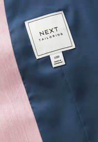 Sezione di tessuto blu navy e rosa chiaro con un'etichetta bianca che riporta la scritta "NEXT TAILORING", taglia 40R, e "FABBRICATO IN MYANMAR". Finitura strutturata.
