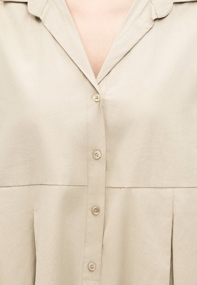 Εκρού ένδυμα με κουμπιά, γιακά και κάθετες πιέτες κάτω από τα κουμπιά, εμφανιζόμενο στο άνω μέρος του κορμού.